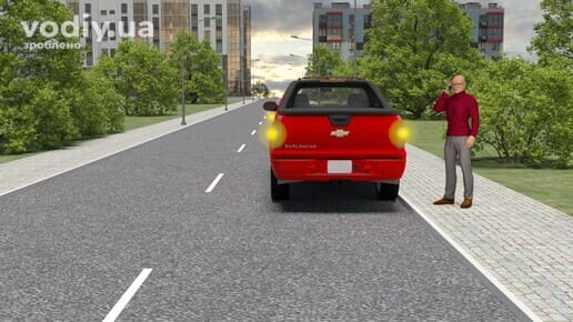 Червоний пікап з увімкненою аварійною світловою сигналізацією біля краю проїзної частини, водій розмовляє по телефону поруч із авто.