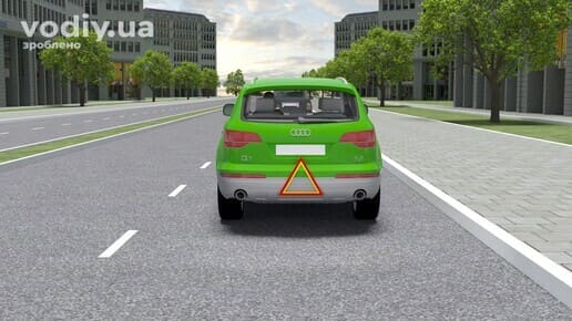 Зелений автомобіль рухається дорогою зі знаком аварійної зупинки, закріпленим на задньому бампері.