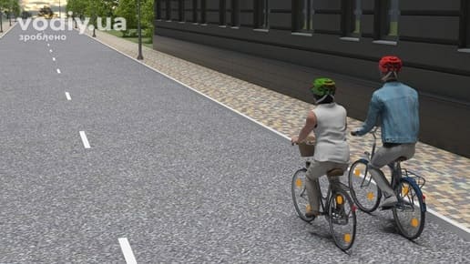 Двоє велосипедистів у шоломах рухаються поруч в один ряд по проїзній частині вздовж тротуару, порожня дорога в населеному пункті.