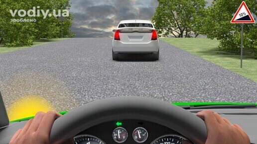 Білий автомобіль попереду на підйомі дороги, дорожній знак 1.6 «Крутий підйом», увімкнений лівий покажчик повороту в авто водія.