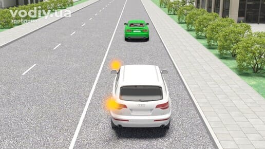 Білий автомобіль з увімкненим лівим покажчиком повороту позаду зеленого авто на дорозі з двома зустрічними смугами. Обгін на дорозі з трьома смугами.